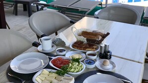 Uma mesa de jantar com diversos alimentos e bebidas, com vista para uma área de piscina com espreguiçadeiras e guarda-sóis.