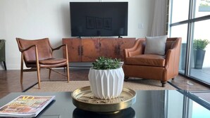 A modern living room with a glass coffee table, leather armchairs, a white vase with a plant, and a flat-screen TV on a wooden cabinet.