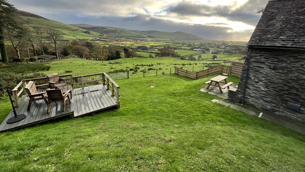A scenic outdoor area with a wooden deck and picnic table, surrounded by lush green fields and hills in the background. - Converted Barn. Dogs welcome. Dog play field. Walk to local pub. (Tywyn)