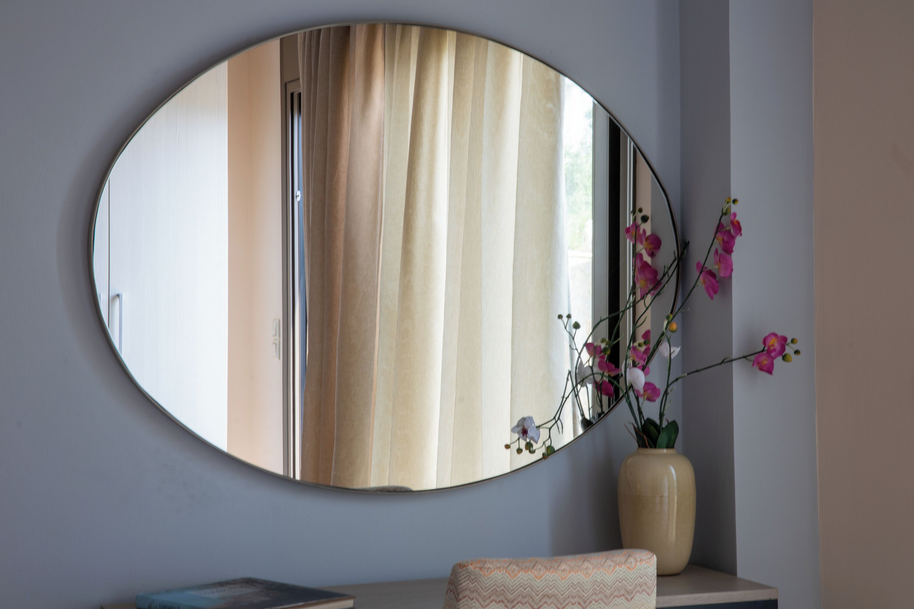 Una habitación con un espejo ovalado grande que refleja una ventana con cortinas, un jarrón con flores y una pequeña mesa con un libro.