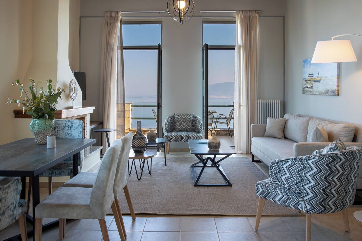 A modern living room with a sofa, chairs, a dining table, and a view through large windows.