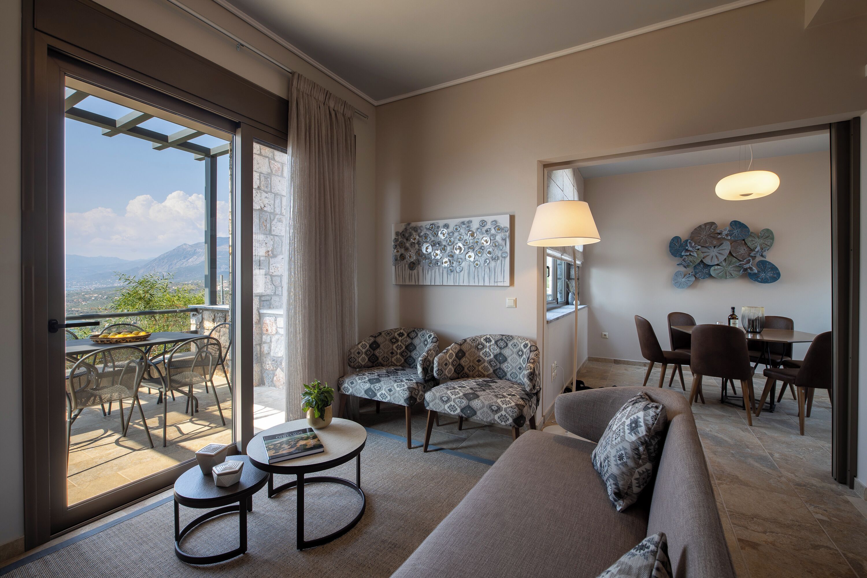 A modern living room with a sofa, armchairs, a dining table, and a balcony with a view of the mountains.