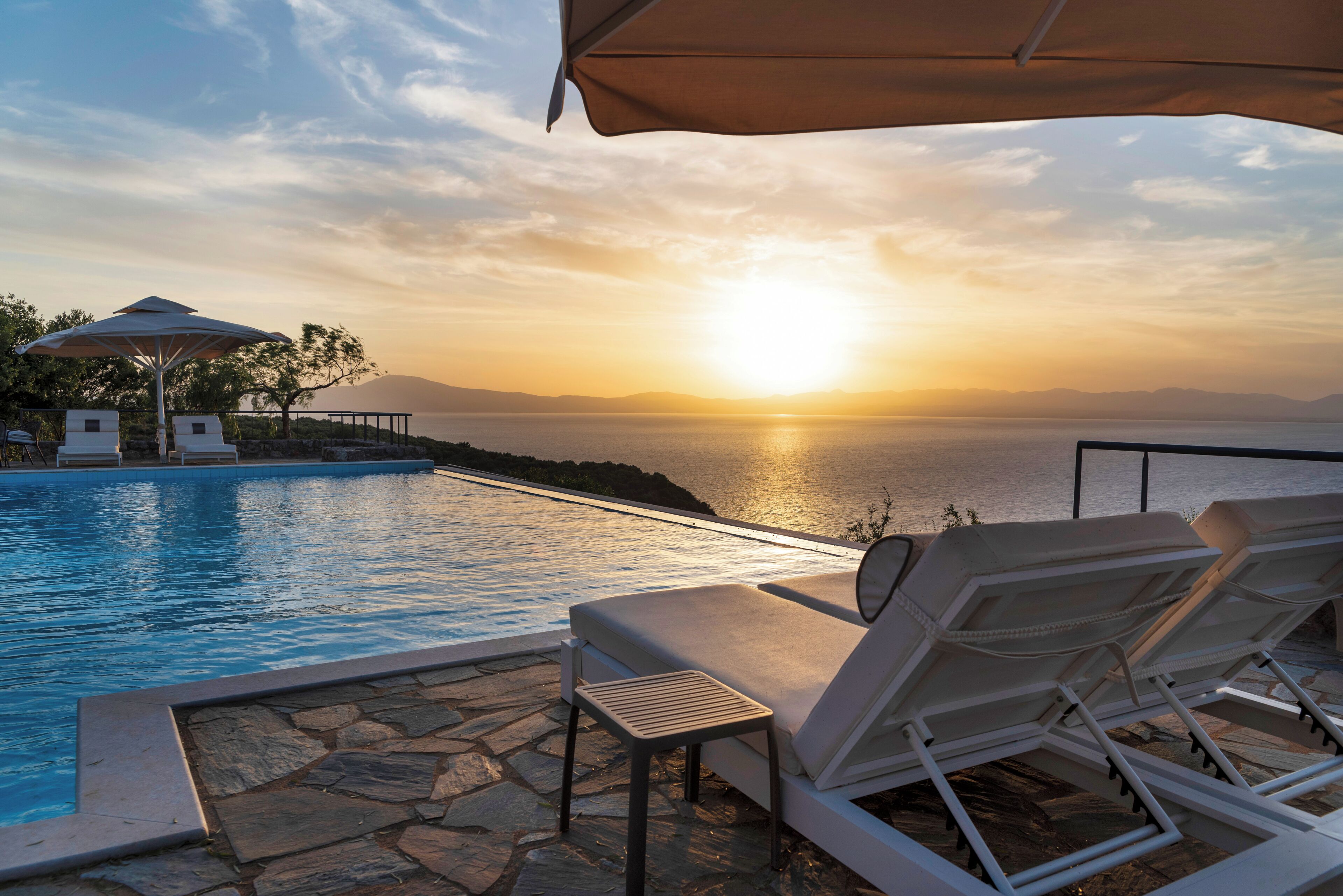 Área de piscina com espreguiçadeiras e guarda-sóis, com vista para um corpo d'água e um pôr do sol.