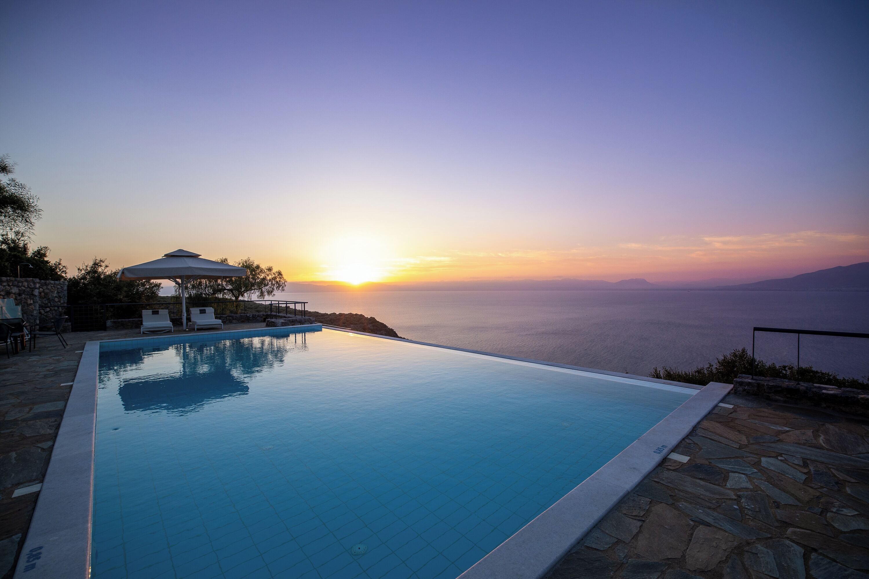 Una piscina con vista al atardecer, suelo de piedra y sillas de descanso.