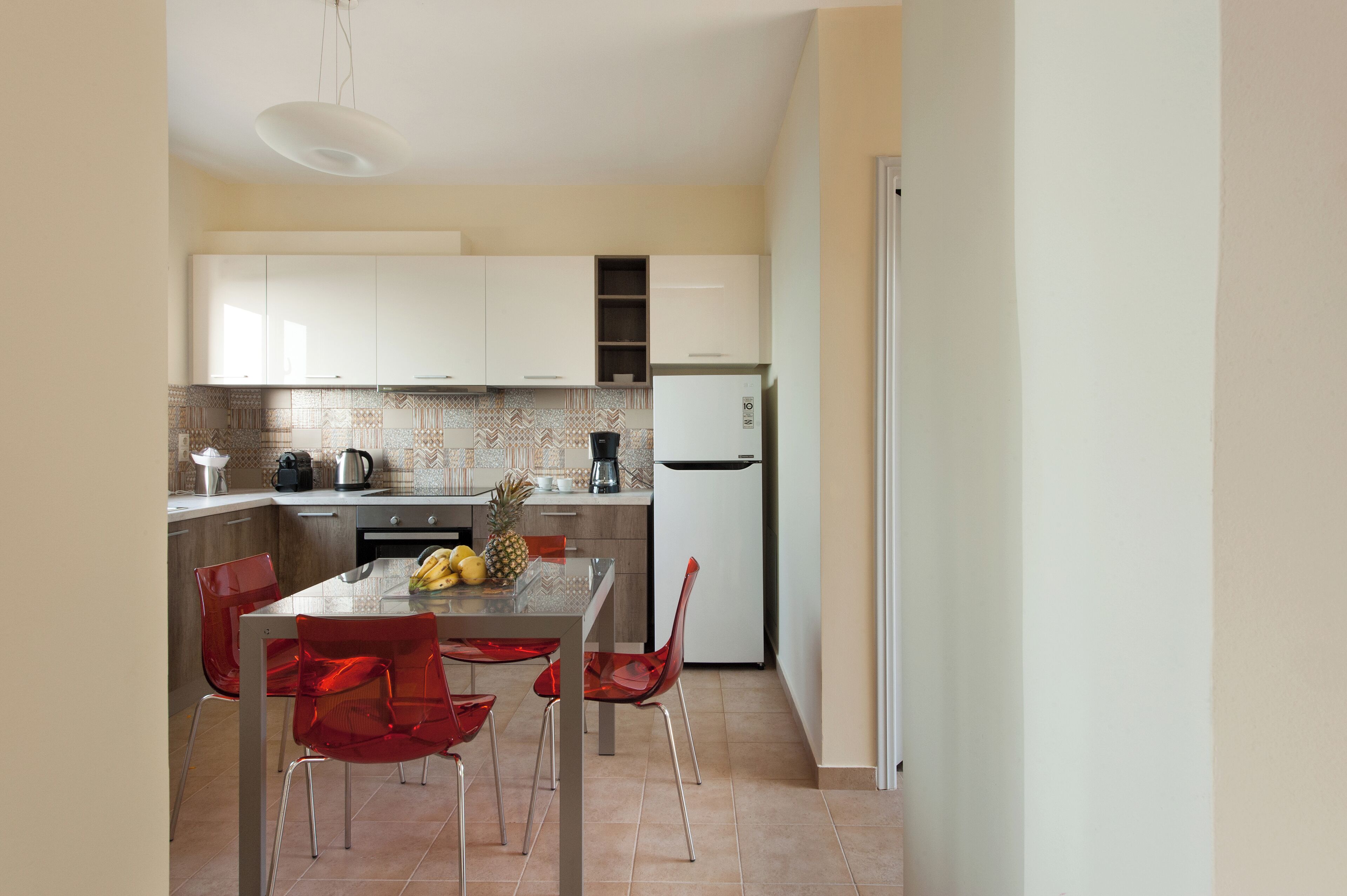 A modern kitchen with white cabinets, a stainless steel refrigerator, and a dining area with a glass table and red chairs.
