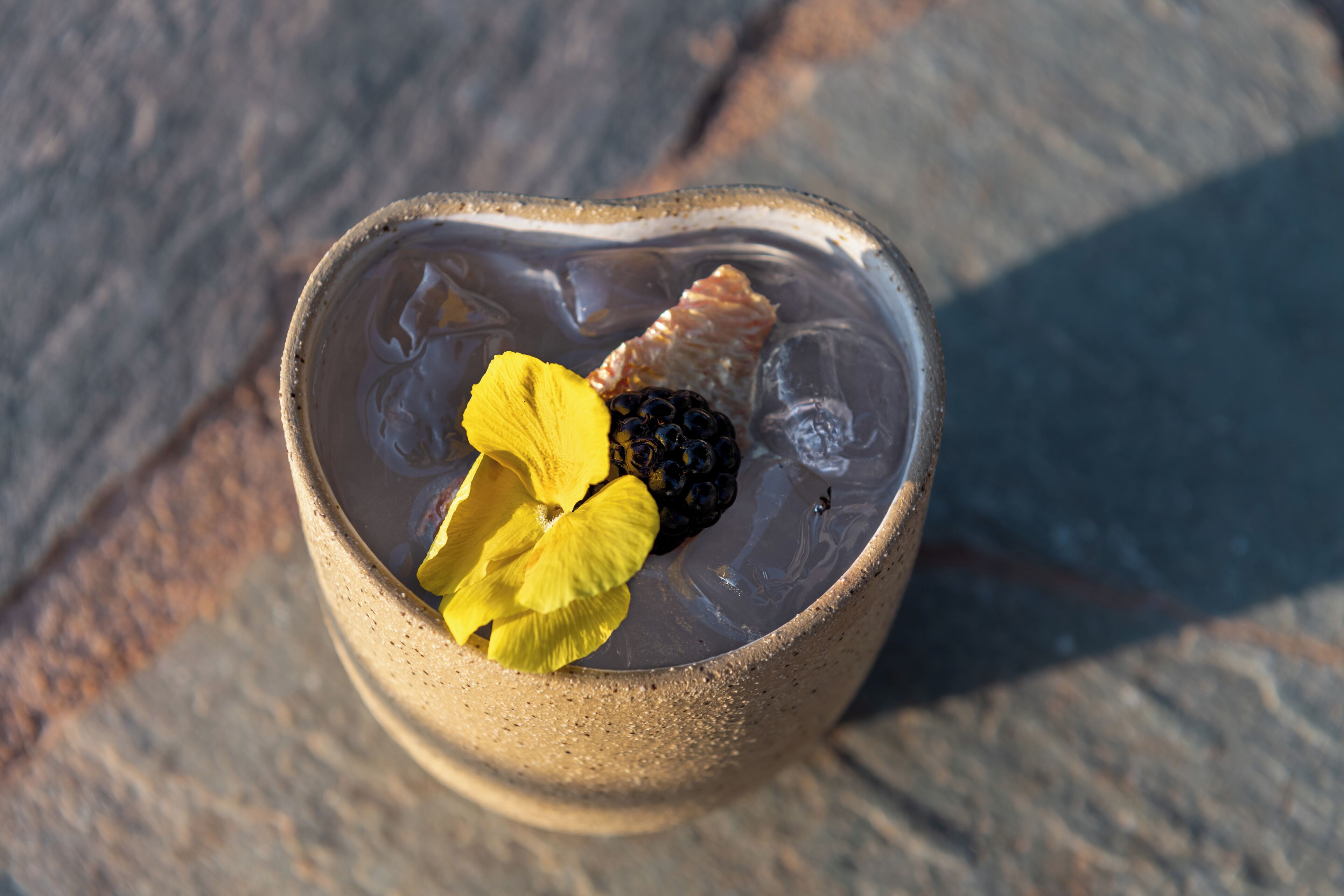 A clear glass with ice cubes, a yellow flower, and a blackberry on a stone surface.
