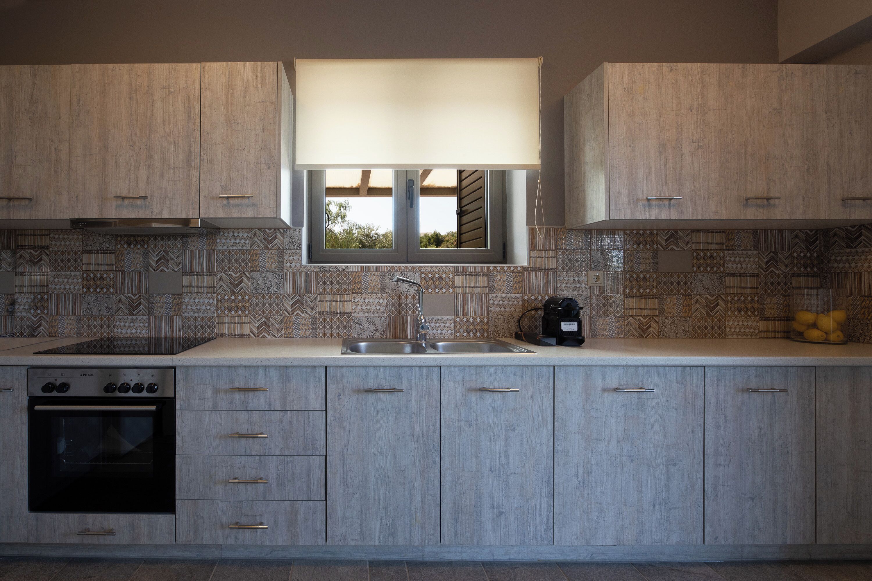 A modern kitchen with light wood cabinets, a patterned backsplash, a built-in oven, and a window above the sink.