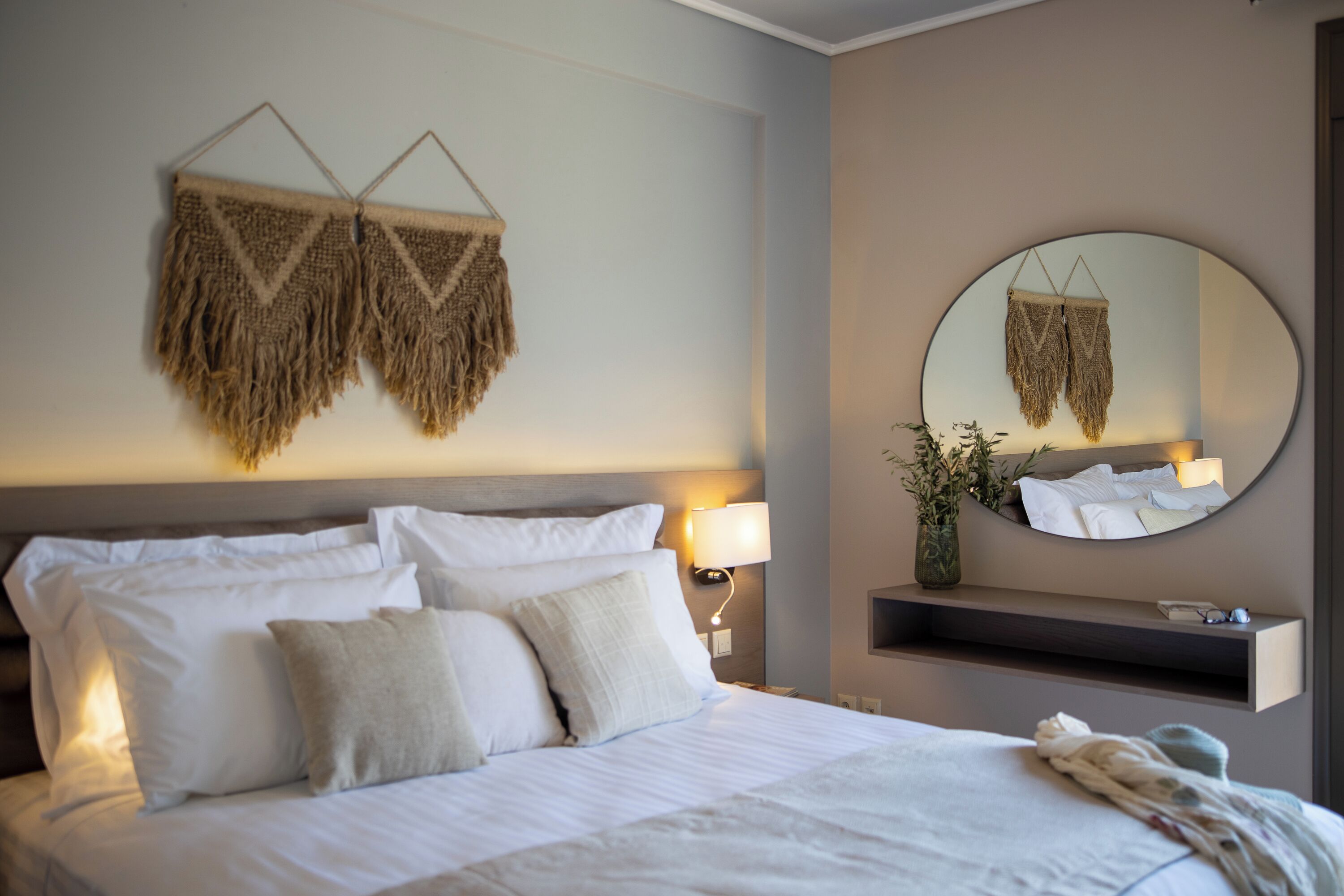 A neatly made bed with white linens and pillows, a bedside lamp, a wall-mounted mirror, and a small shelf with a vase and a plant.