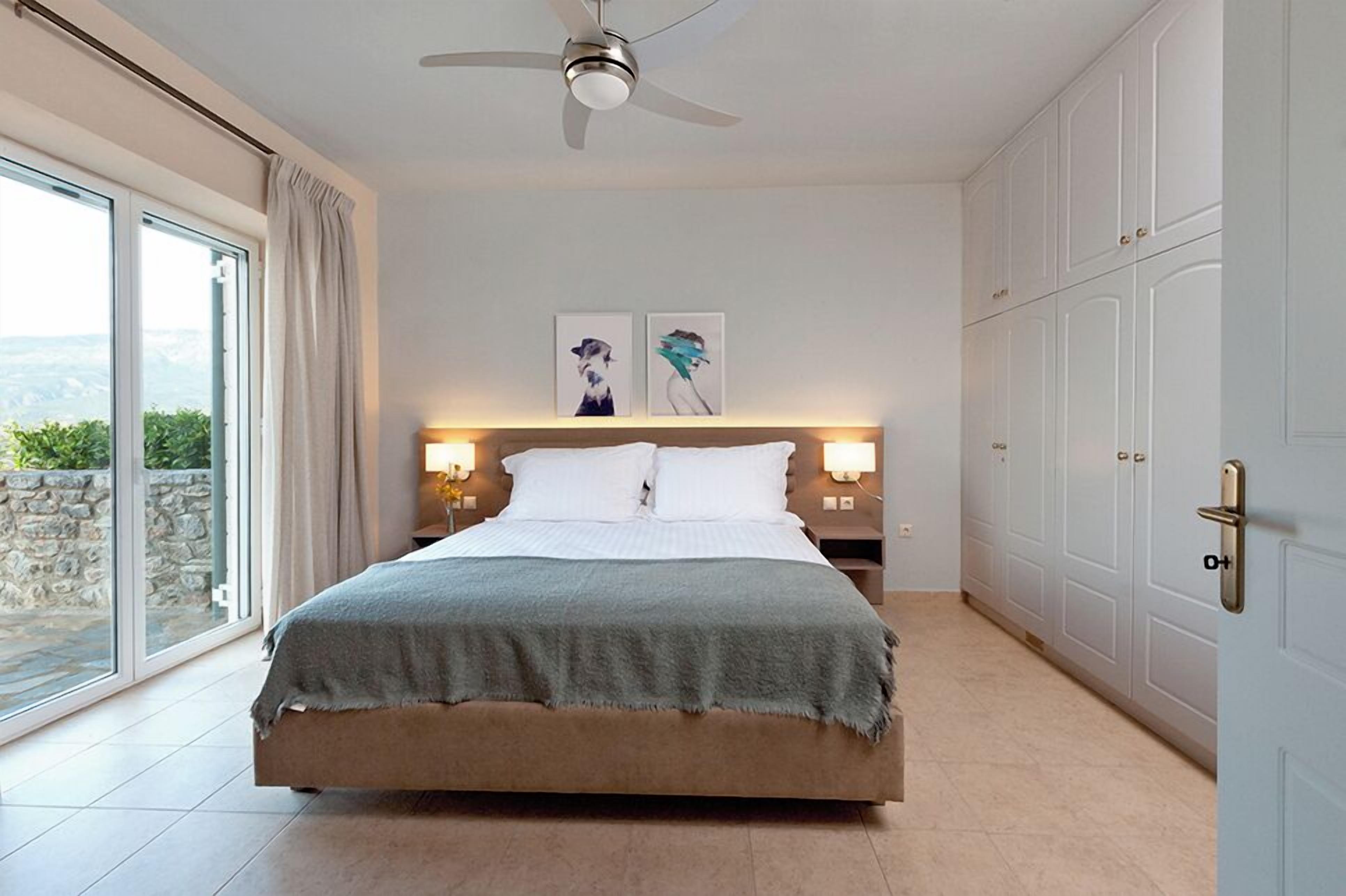 Un dormitorio con cama, ventilador de techo, mesita de noche, lámparas de pared y puerta corrediza que da a un balcón.