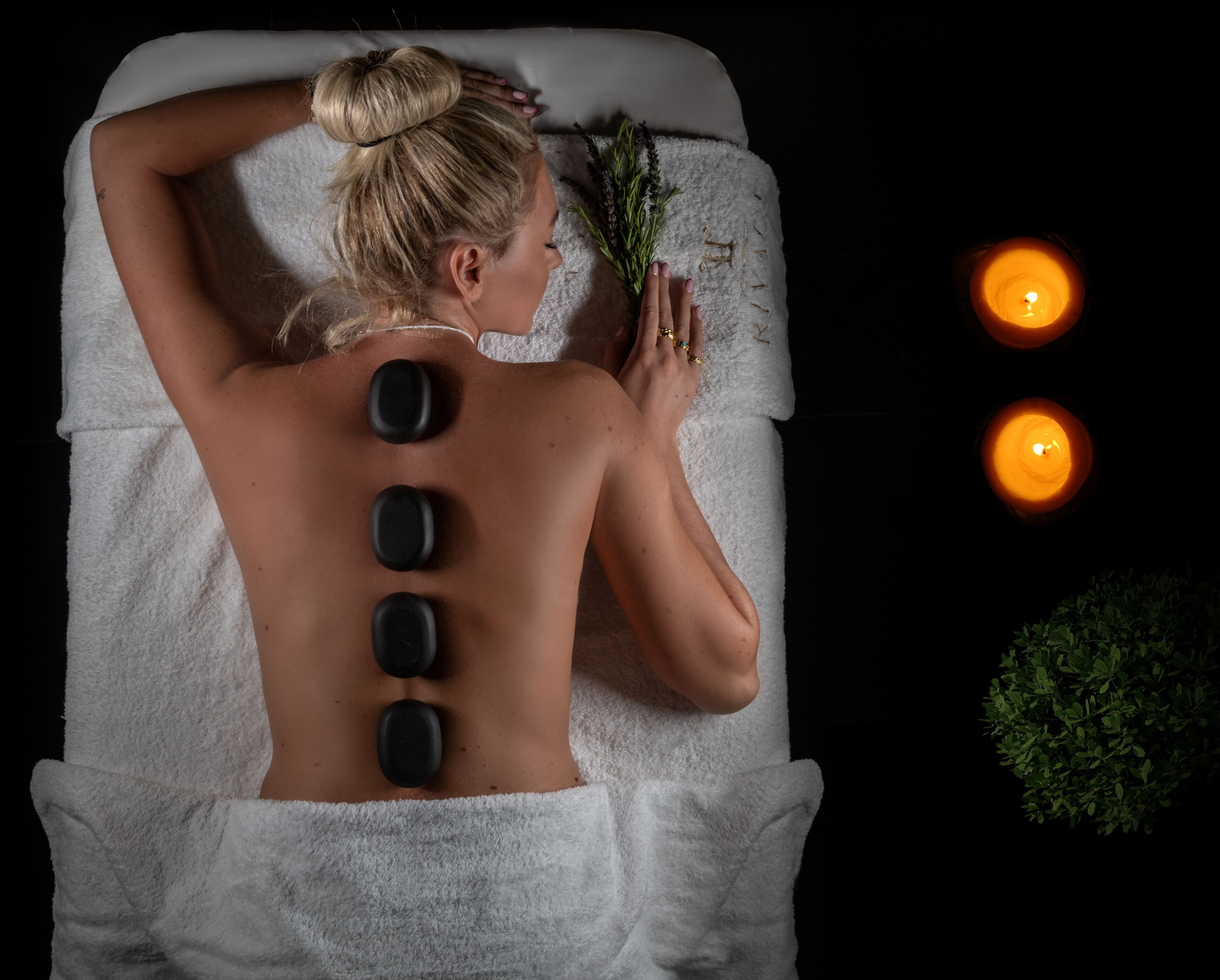 A person receiving a back treatment with stones, candles, and a plant in a dimly lit room.