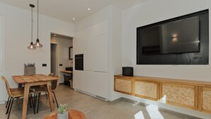 A modern kitchen with white cabinets, a wooden dining table, and a large flat-screen TV mounted on the wall.