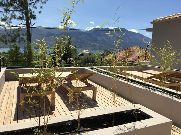 Terraço no telhado com móveis de madeira e plantas de bambu, com vista para uma paisagem montanhosa e um corpo d'água.