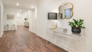 Un pasillo con piso de madera, una mesa consola de vidrio transparente con una lámpara, una planta en maceta y un espejo montado en la pared.