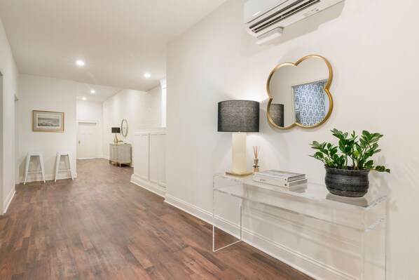 A hallway with wooden flooring, a clear glass console table with a lamp, a potted plant, and a wall-mounted mirror.