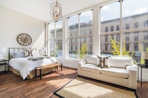 Makuuhuoneessa on suuri ikkuna, sänky, penkki, sohva ja seinäkello.