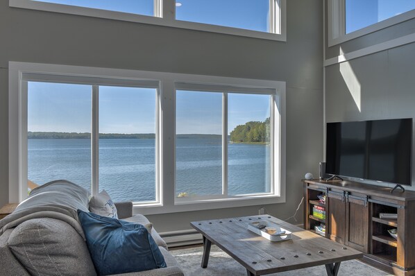 A living room with a large window overlooking a lake, a television, and a wooden coffee table. - Just steps to the water with beautiful sunsets! (Waldoboro)