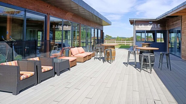 An outdoor seating area with wicker furniture and tables, a wooden deck, and glass walls offering views of the surrounding landscape. - House La Houle Erquy *Pool* *Beach at 800m* (Erquy)