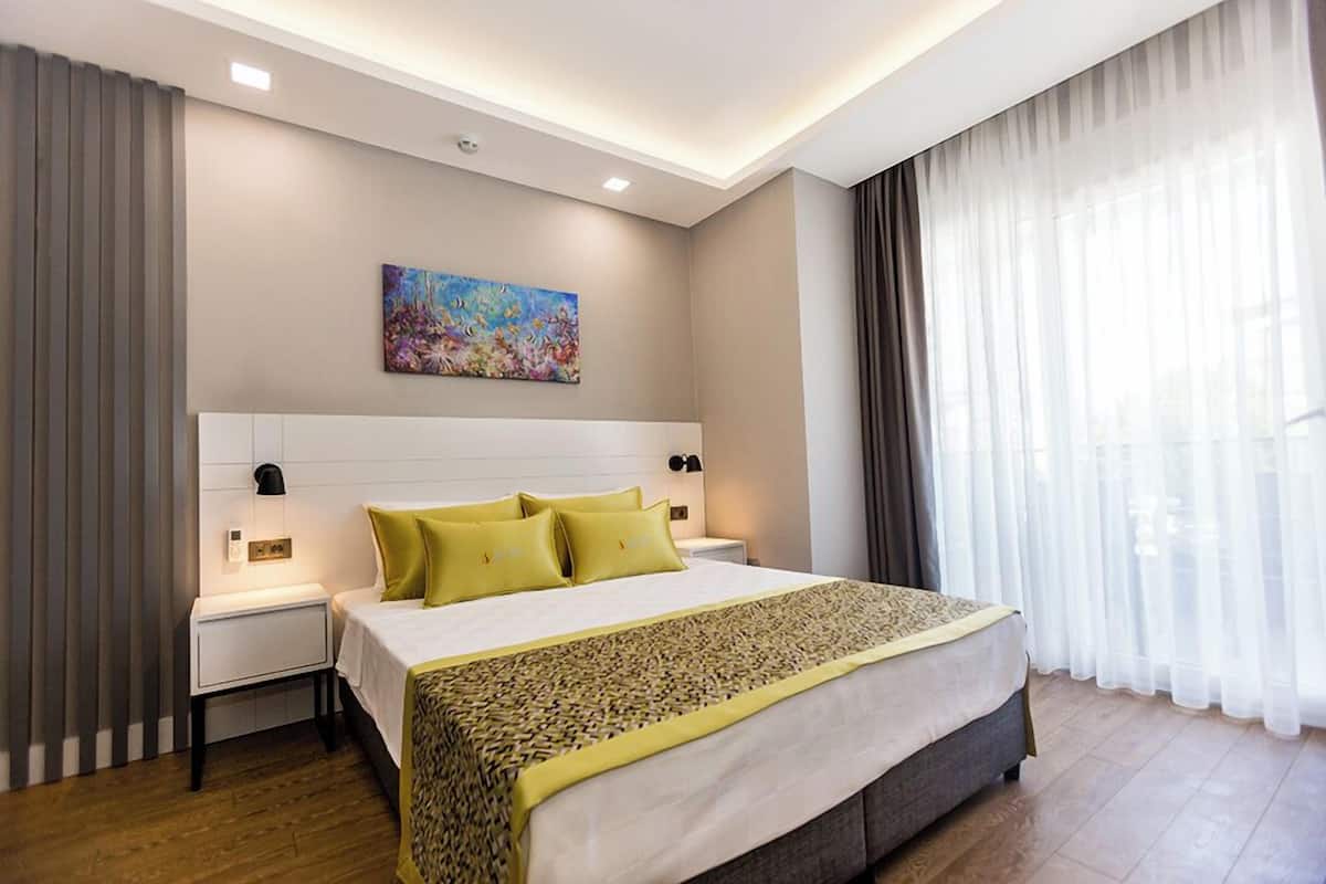 A modern hotel room with a large bed, a nightstand, a wall-mounted lamp, a painting, and sheer curtains.