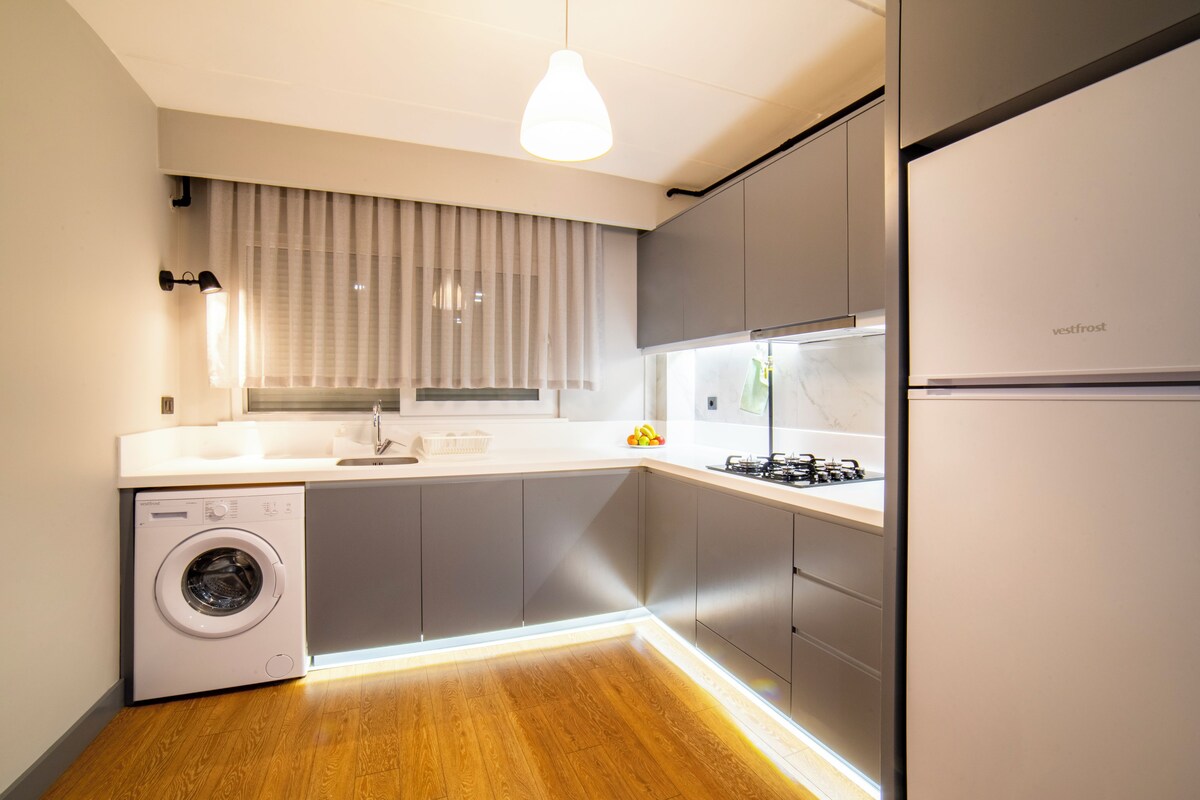 Una cocina moderna con lavadora integrada, armarios grises, un refrigerador de acero inoxidable y una lámpara colgante.