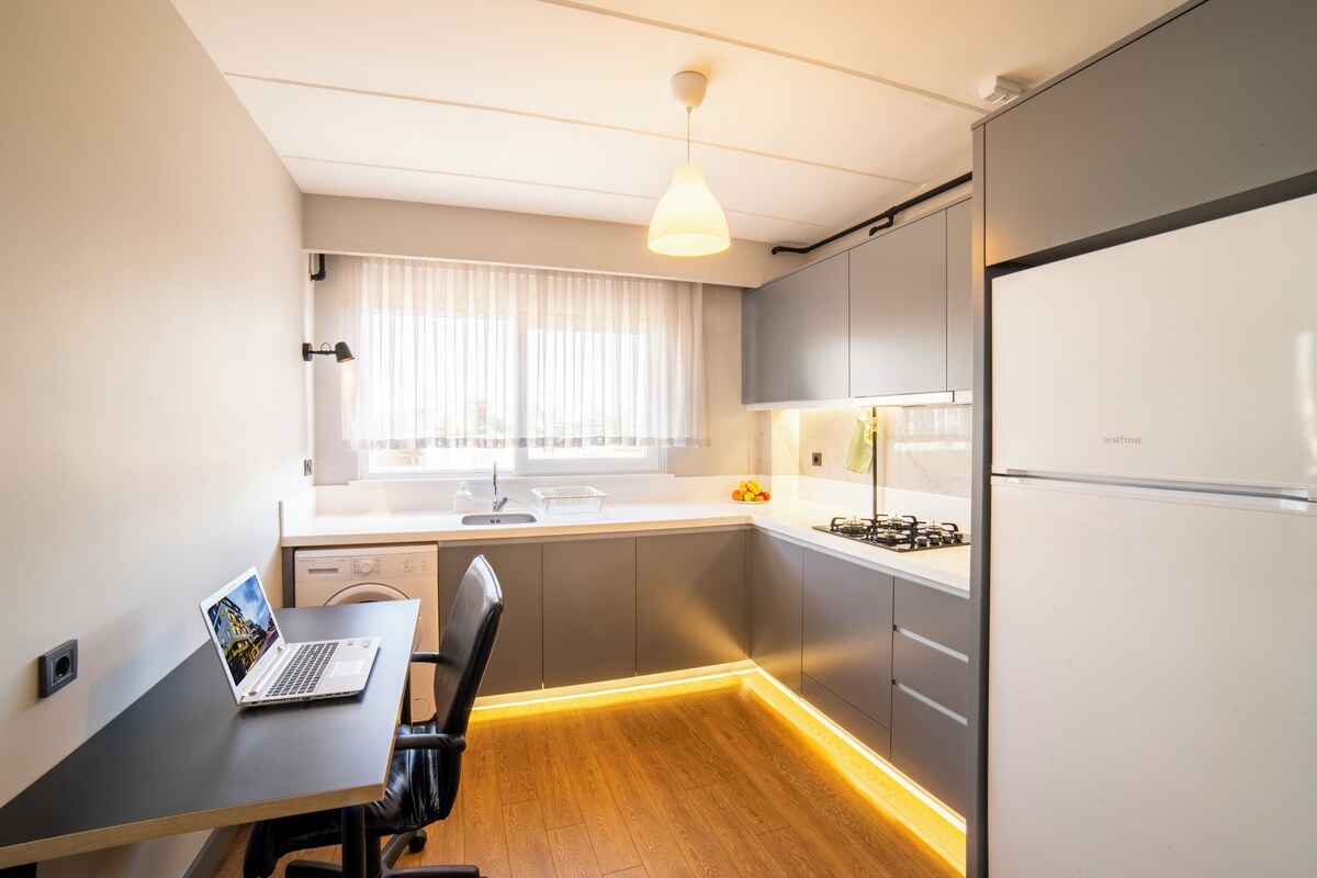 Una cocina compacta con una laptop sobre una mesa, un refrigerador y una ventana con cortinas.