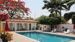 Una piscina con sillones de playa y sombrillas, rodeada de palmeras y un edificio con techo de tejas.
