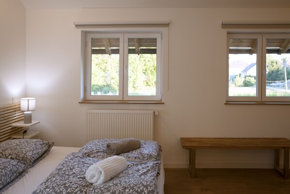 A bedroom with a bed, a bench, a nightstand with a lamp, and two windows with a view of greenery.