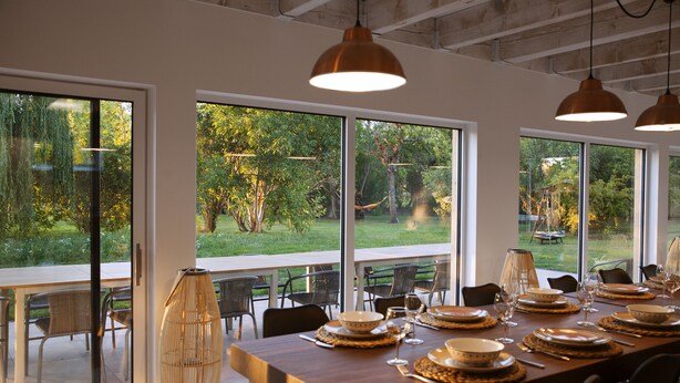 A dining area with a long table set for a meal, large windows offering a view of the outdoors, and pendant lighting.