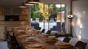 A dining area with a long wooden table set for a meal, overhead pendant lighting, and a view of a garden through large sliding doors.