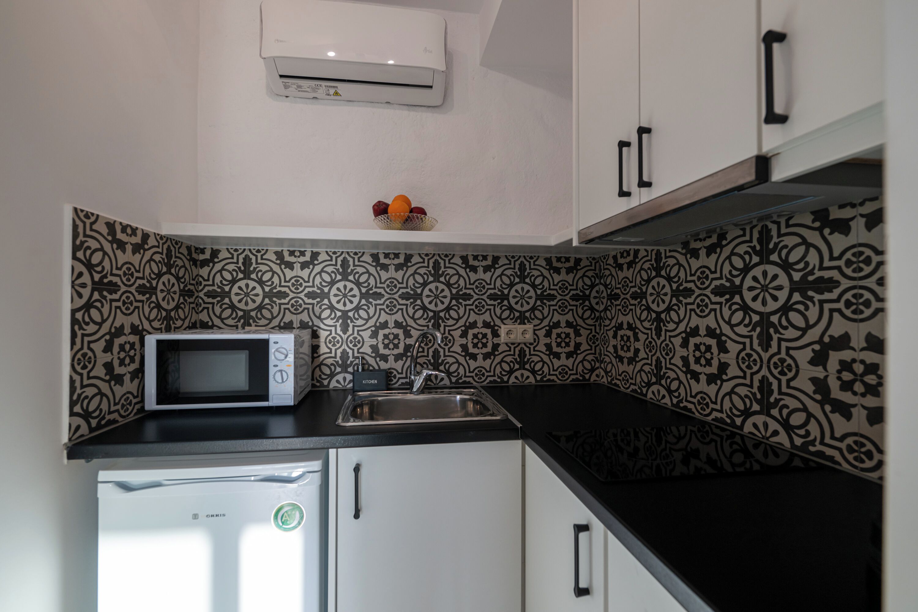 A compact kitchen with a microwave, refrigerator, and sink. The backsplash features a black and white patterned tile design.