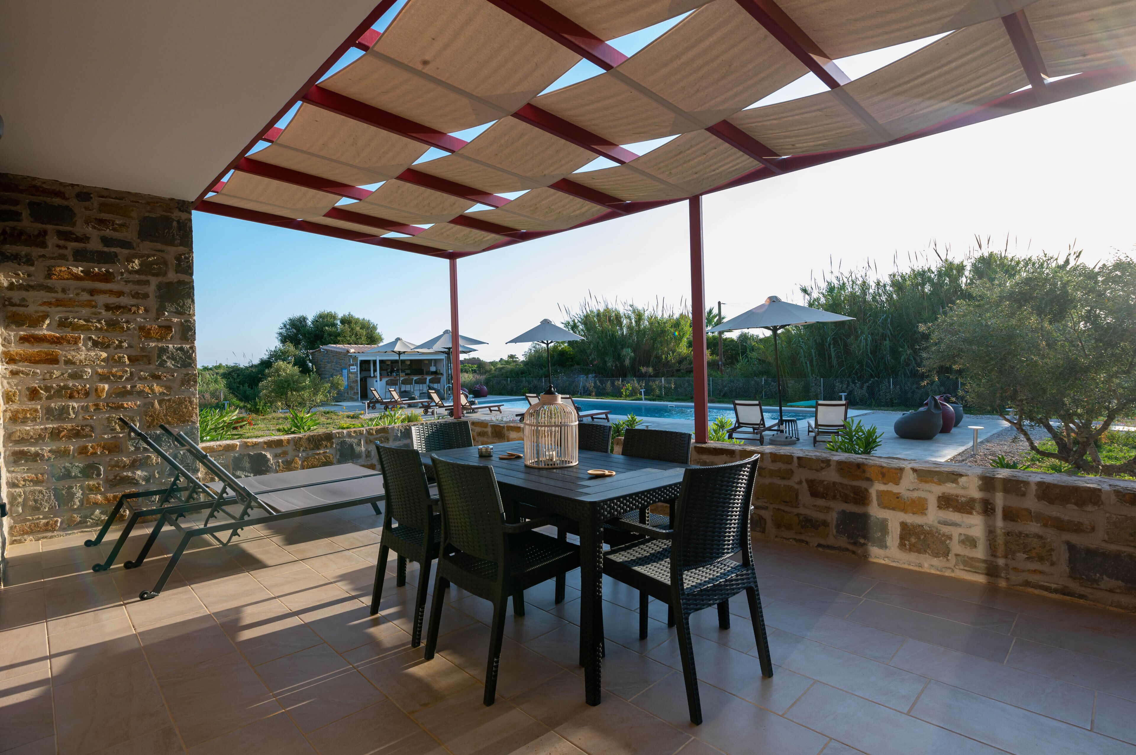 Un área de estar al aire libre con techo, vista a la piscina, paredes de piedra y mobiliario exterior.
