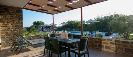 Un área de estar al aire libre con techo, vista a la piscina, paredes de piedra y mobiliario exterior.