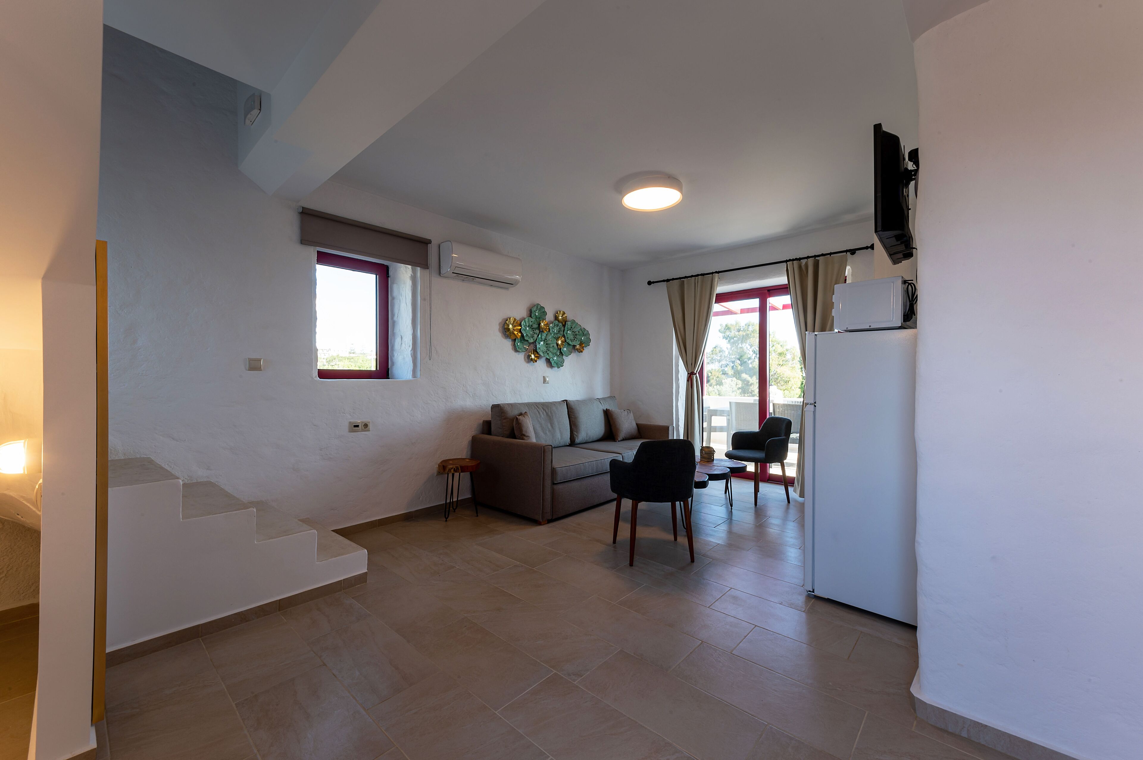 A spacious living area with a sofa, a chair, a small table, and a refrigerator. There is a staircase leading to another level, and a window with curtains.