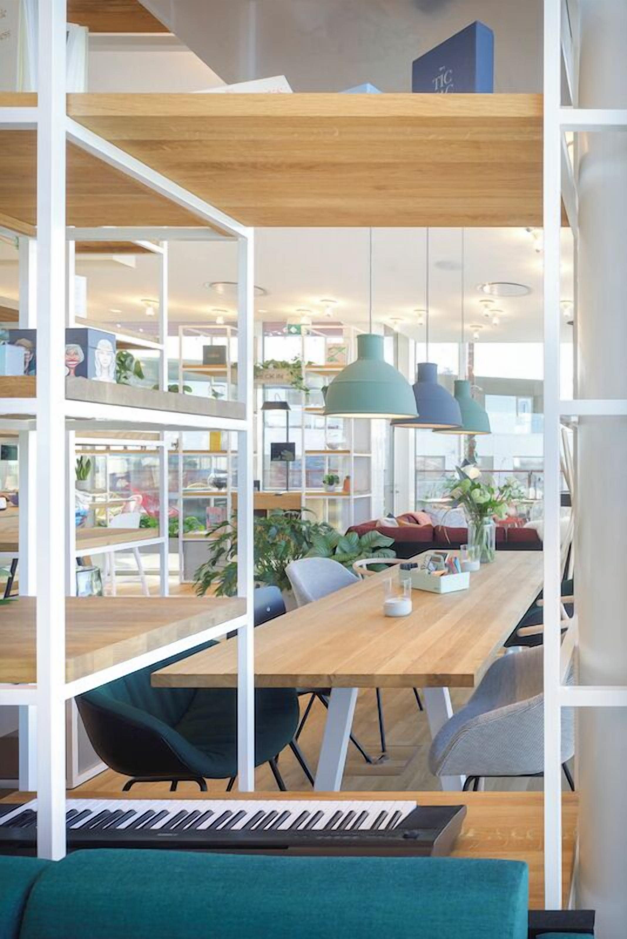 A modern workspace with a wooden table, chairs, a keyboard, shelves with plants, and pendant lights.