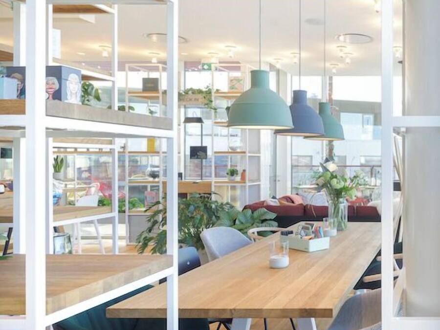 Espaço de trabalho moderno com mesa de madeira, cadeiras, teclado, prateleiras com plantas e luminárias pendentes.