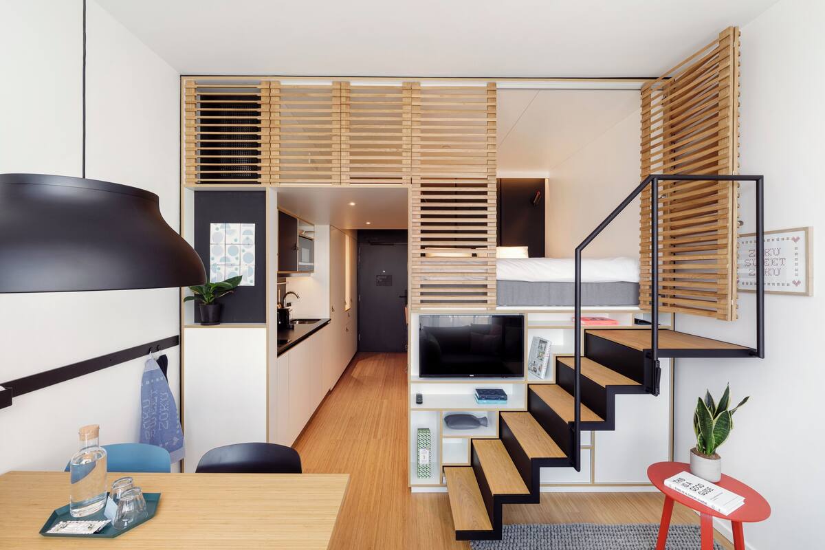 Een kleine hotelkamer met een bed, een televisie, een bureau, een stoel, een tafeltje, een rode kruk, een plant en een trap.
