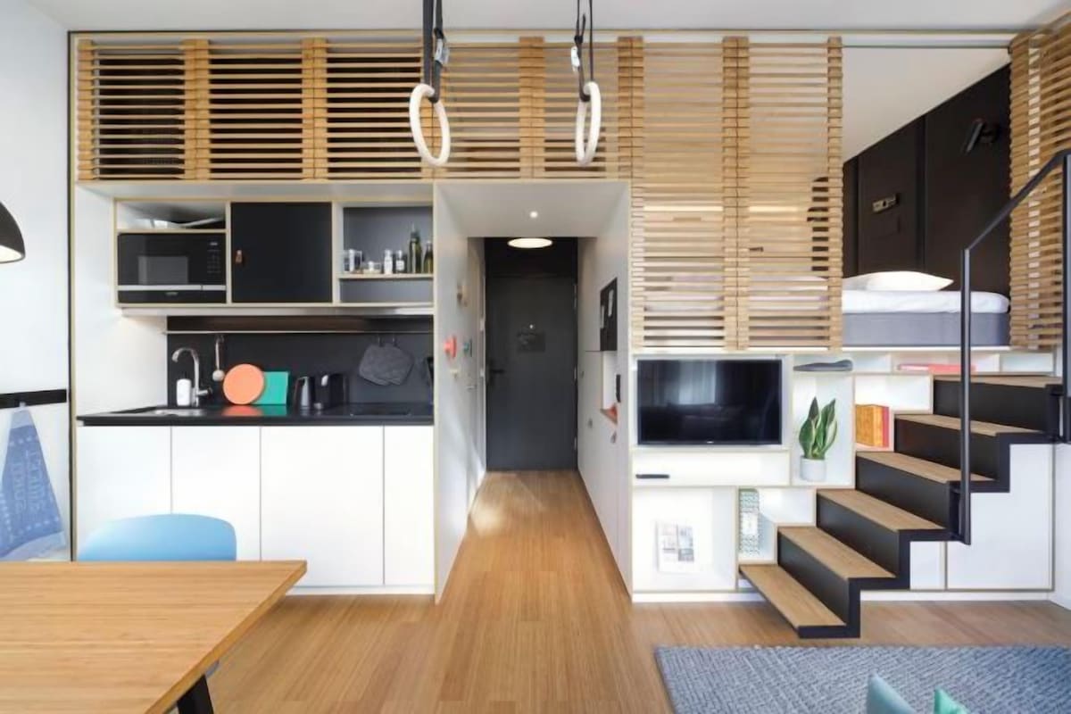 Una cocina moderna con electrodomésticos integrados, un comedor con mesa de madera y una escalera que sube a un dormitorio tipo loft.
