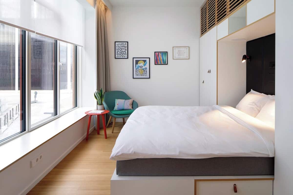 Een moderne hotelkamer met een groot raam, een bed met witte lakens, een klein rode krukje, een blauwgroene stoel en meerdere lijstwerken aan de muur.