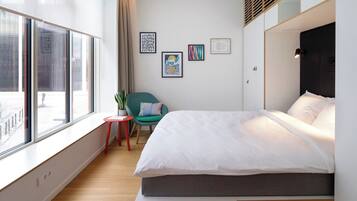 A modern hotel room with a large window, a bed with white linens, a small red stool, a teal chair, and several framed artworks on the wall.