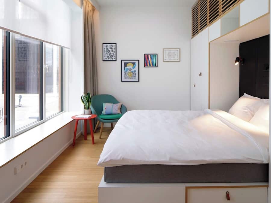 A modern hotel room with a large window, a bed with white linens, a small red stool, a teal chair, and several framed artworks on the wall.