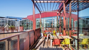Een terras met tafels en stoelen, een pergola met lichtslingers en grote glasramen.