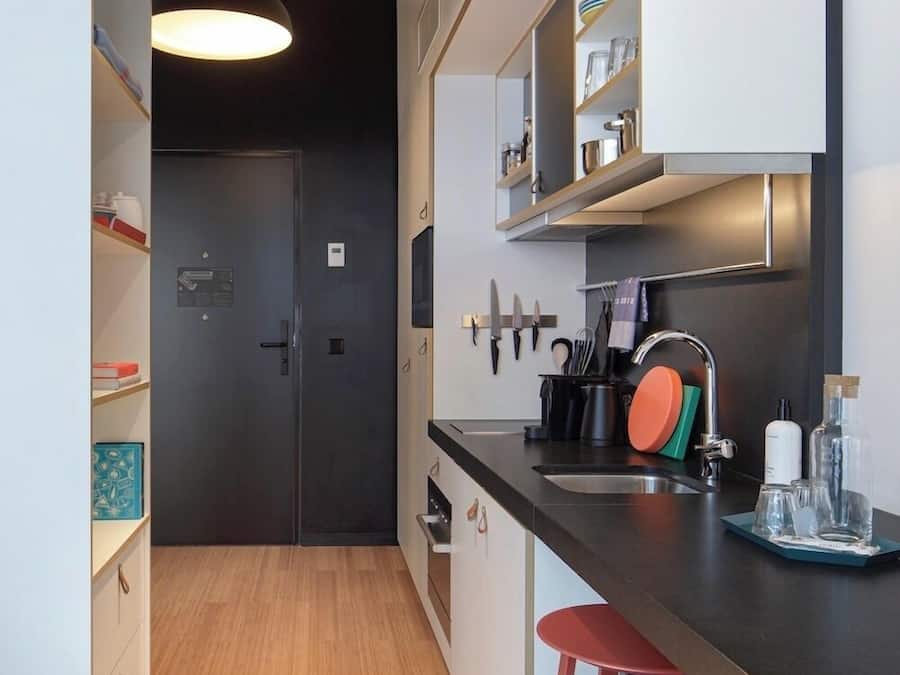 A modern kitchen with a dark door, wooden flooring, a bar area with stools, and a hanging light fixture.