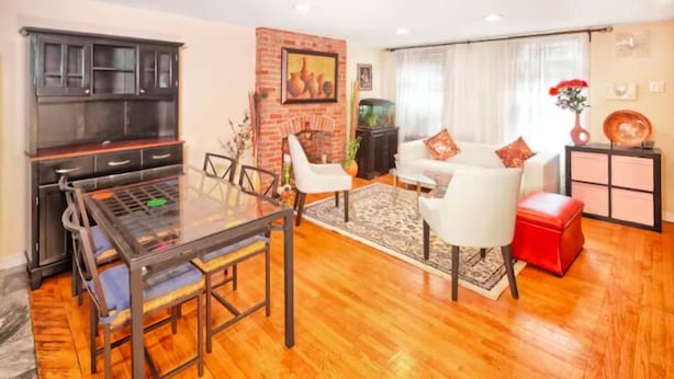 Un salon comprenant une table à manger avec un plateau en verre, une cheminée, un ensemble de canapés et un pouf rouge.