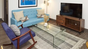 A living room with a blue sofa, a wooden TV stand, a glass coffee table, and a patterned rug. - Luxury Building 3BR/2BA! Prime Downtown Mass Ave! (Indianapolis)