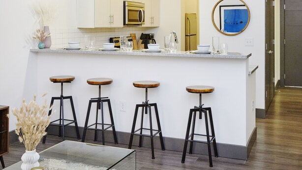 A modern kitchen with white cabinets, a central island with stools, and a dining table with a vase of dried flowers. - Luxury Building 3BR/2BA! Prime Downtown Mass Ave! (Indianapolis)