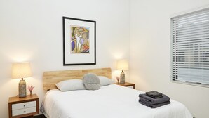 A neatly made bed with white linens, two bedside lamps, a framed artwork above the bed, and a window with blinds. - Luxury Building 3BR/2BA! Prime Downtown Mass Ave! (Indianapolis)