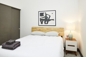 A neatly made bed with white bedding and two pillows, a wooden headboard, a framed abstract artwork, a bedside lamp, and a small vase with flowers on a nightstand. - Luxury Building 3BR/2BA! Prime Downtown Mass Ave! (Indianapolis)