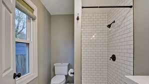 Et badeværelse med toilet, brusebad med hvide fliser og et vindue med rullegardiner.