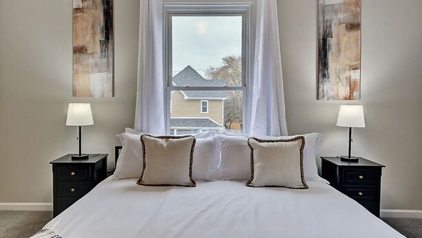 Une chambre avec un grand lit, deux tables de chevet avec des lampes, une fenêtre avec des rideaux et des œuvres d’art abstraites accrochées au mur.