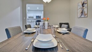 Un coin repas avec une table en bois dressée pour quatre personnes, une cuisine moderne en arrière-plan et un luminaire contemporain.