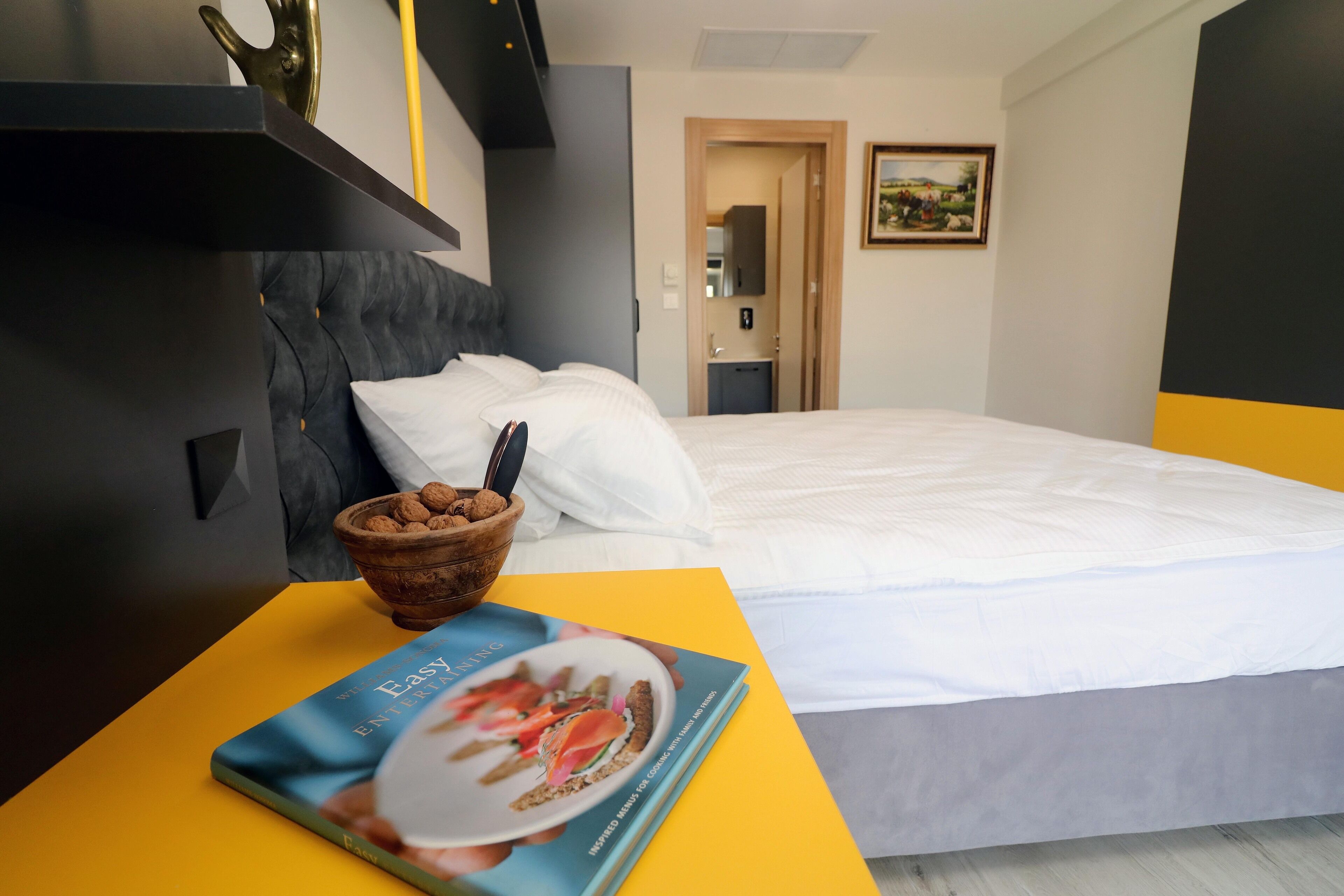 Quarto de hotel com uma cama, uma mesa de cabeceira com uma tigela de lanches e um livro intitulado "Easy Entertaining".